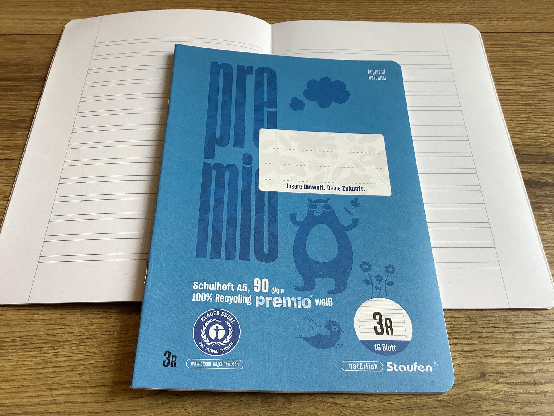 kleines Schulheft mit blauen Cover, einmal geschlossen, darunter einmal aufgeschlagen mit den „Schlauch-Linien“ fürs dritte Schuljahr