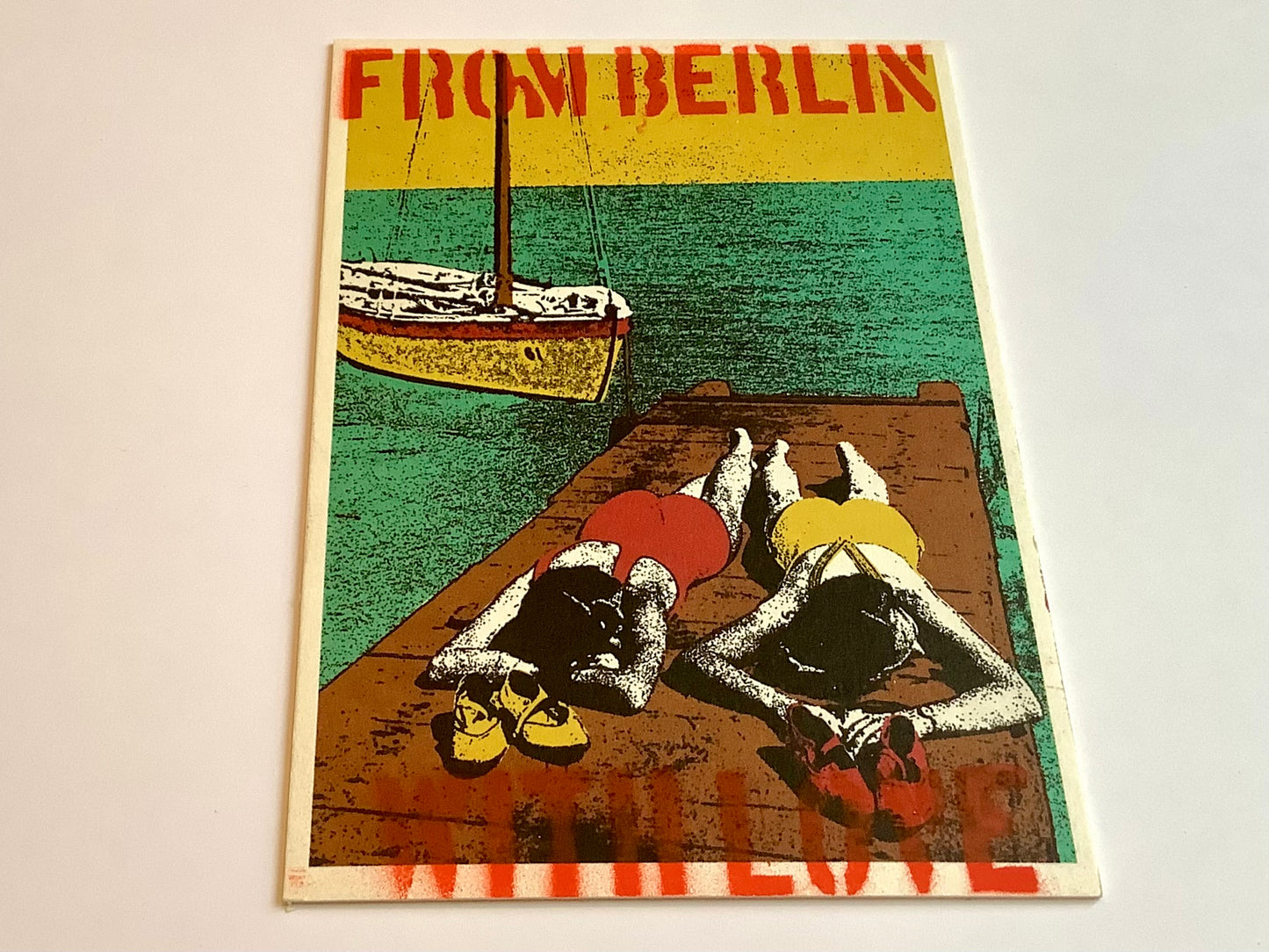 Die illustrierte Postkarte zeigt 2 Frauen im 20er-Jahre Badeanzug auf dem Bauch liegend auf einem Steg am Wasser, Boot im Hintergrund. Der Text oben und unten: From Berlin with love.
