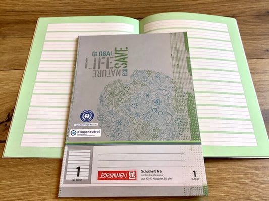 Schulheft A5, aufgeschlagen mit Lineatur 1 vor grünem Kontrasthintergrund, Cover grau mit blaugrüner Weltkugel