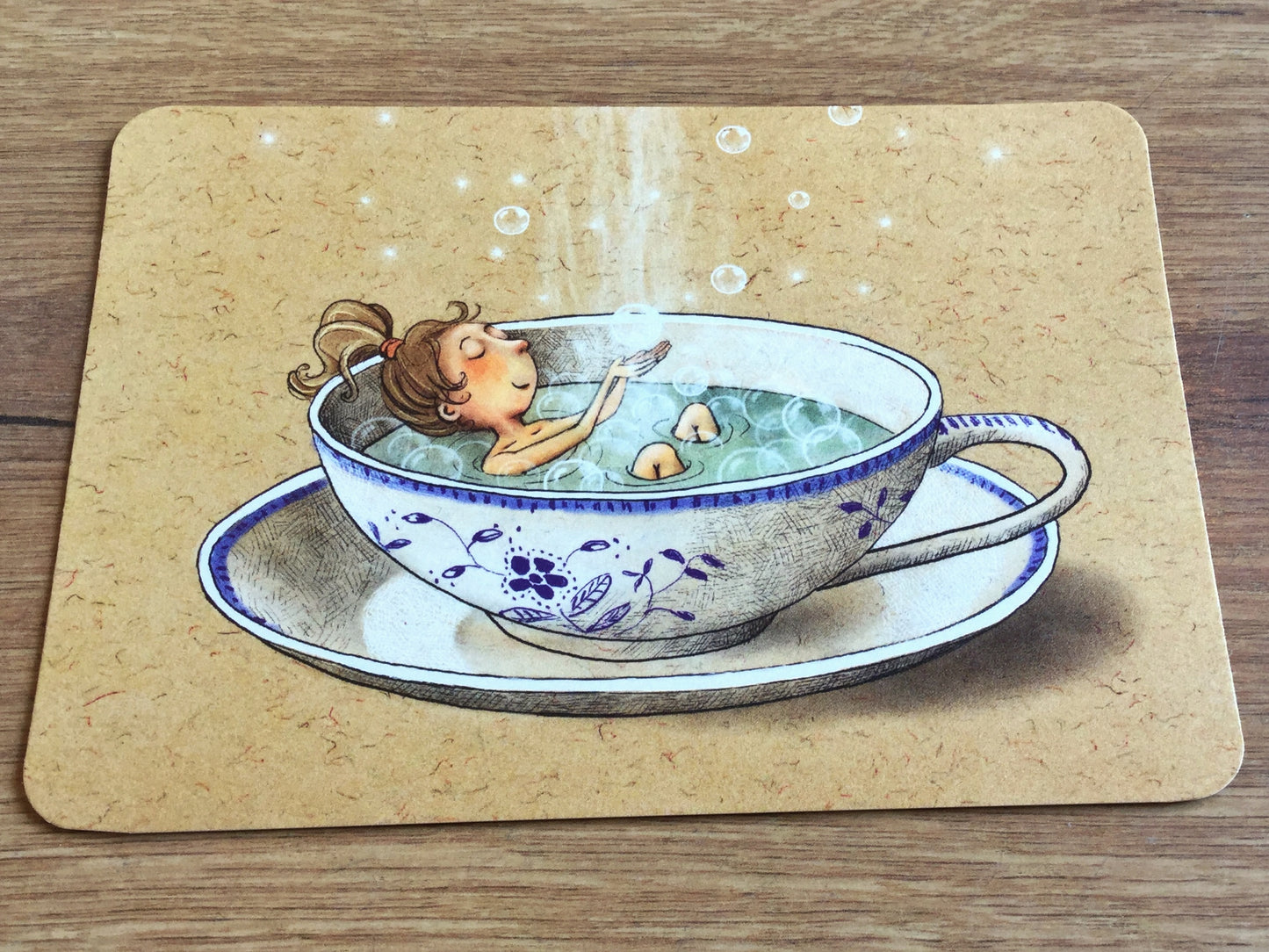 Die Postkarte zeigt eine blauweiß-geblümte Tasse mit dampfendem Tee, in dem ein Mädchen badet und mit Seifenblasen spielt. Gutrath-Verlag. 
Erhältlich bei Polly Paper - Umweltfreundliche Schreibwaren in Berlin Mitte