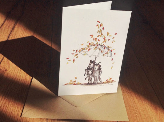Klappkarte Herbst (Inka Erichsen) , ein Zebra und ein Wolf umarmen sich und halten einen großen weißen Schirm über sich, auf den  es bunte Herbstblätter regnet. Darunter ganz klein der Text: irgendwann ist immer Herbst